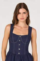 A woman is wearing a sleeveless navy blue dress with wide shoulder straps. The bodice has a square neckline and features vertical and curved white stitching details that form a pattern resembling a fitted, structured design. There are also decorative silver buttons running down the front center of the bodice. The skirt portion is gathered at the waist, creating a slightly flared silhouette.